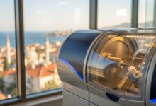 A close-up, photorealistic shot of a modern dental CAD/CAM machine creating a white ceramic crown, with a stunning, blurred background of a sunny Turkish coastal city and the sea, viewed from a clinic window.
