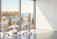 An empty, high-tech dental treatment room with a modern chair in the foreground and a large window offering a soft-focus, sunny view of a beautiful Turkish coastal city and the sea.