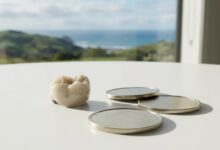 A photorealistic close-up of a porcelain dental crown and New Zealand dollar coins on a clean surface, with the beautiful, blurred coastline of New Zealand visible in the background, symbolizing the cost of dentistry.