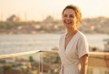 A photorealistic image of a happy woman in her 40s with a brilliant white smile, standing on a hotel balcony with a beautifully blurred view of the Istanbul, Turkey skyline at sunset in the background.