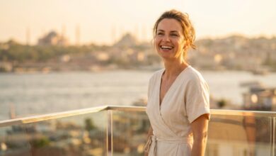 A photorealistic image of a happy woman in her 40s with a brilliant white smile, standing on a hotel balcony with a beautifully blurred view of the Istanbul, Turkey skyline at sunset in the background.
