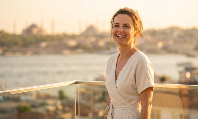 A photorealistic image of a happy woman in her 40s with a brilliant white smile, standing on a hotel balcony with a beautifully blurred view of the Istanbul, Turkey skyline at sunset in the background.