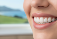 Close-up of a person's natural, healthy, and radiant smile with bright, evenly shaped teeth, indicative of successful cosmetic dentistry. The background is a highly blurred modern dental clinic interior, with a very soft and indistinct hint of green natural landscape, suggesting New Zealand.
