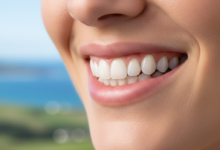 Close-up of a person's mouth showcasing a healthy, radiant smile with perfectly contoured pink gums and bright white teeth. The background is a soft, out-of-focus natural landscape, subtly hinting at New Zealand scenery.