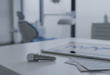 A close-up, photorealistic view of a polished dental implant resting on a modern desk. Beside it are blurred, abstract financial documents with graphs and forms, and a softly out-of-focus clean office or dental clinic in the background. No text is visible.